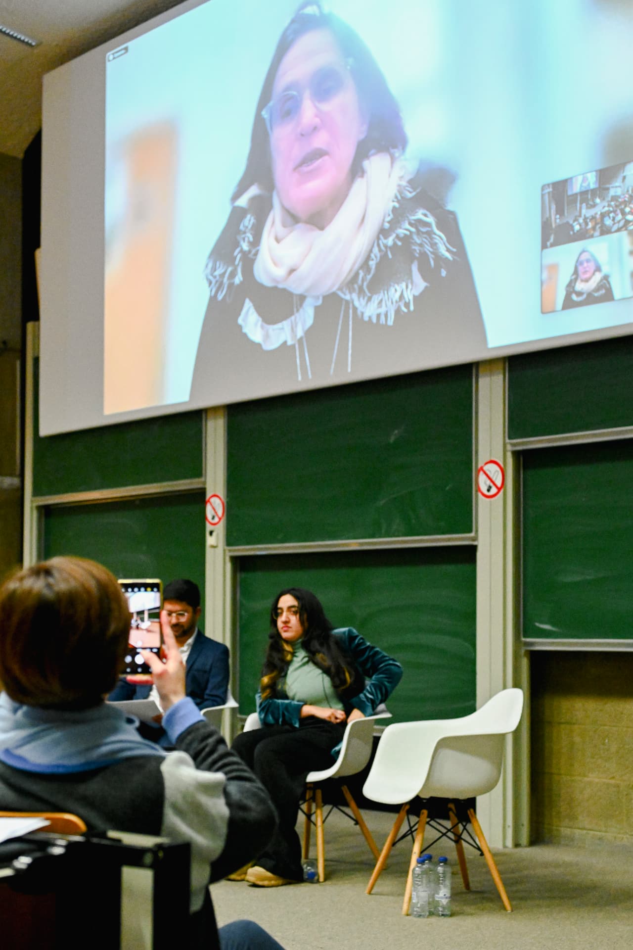 Audience member photographing a woman on stage with a video call projection